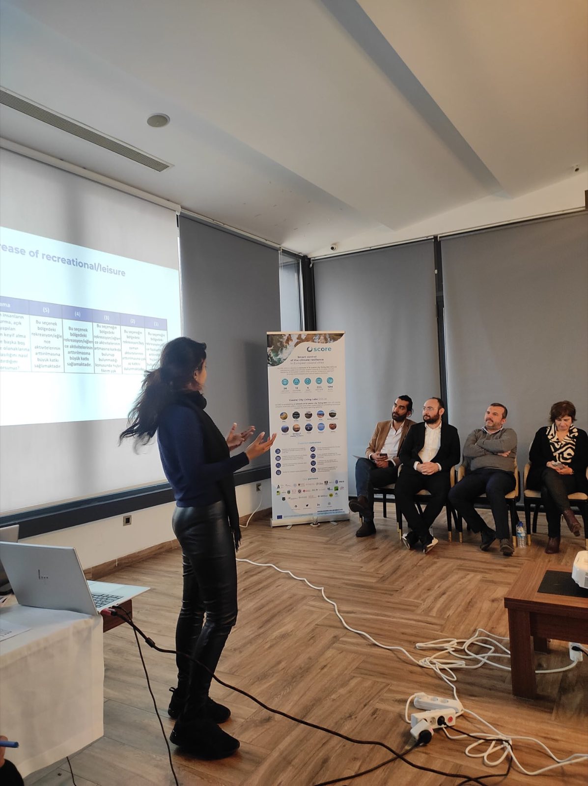 Ananya Tiwari presenting beside a SCORE project banner to a seated panel in Samsun, Turkey